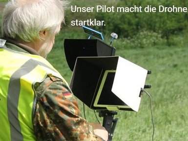 Ein Mann mit weißem Haar trägt eine neongelbe Warnweste mit dem Logo „Kitzrettung Hüttener Berge e. V.“ und blickt auf einen durch einen Sonnenschutz abgeschirmten Monitor. Neben ihm steht eine weitere Person in einer ländlichen Umgebung.